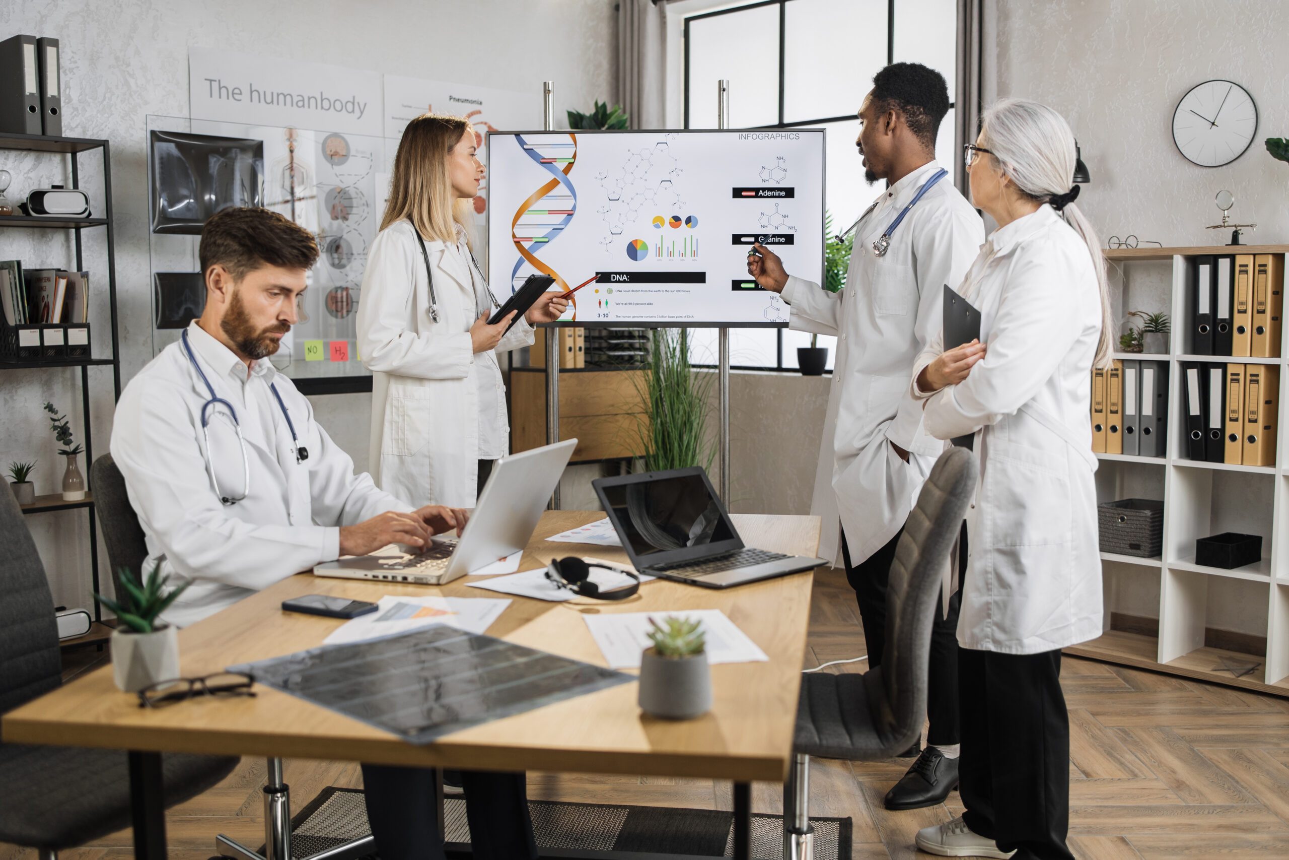 Doctors with laptop having sesion in lab boardroom Group of multiethnic concentrated doctors standing near screen during working sesion in lab boardroom. Caucasian male working on laptop while team of colleagues talking about genetic engineering.